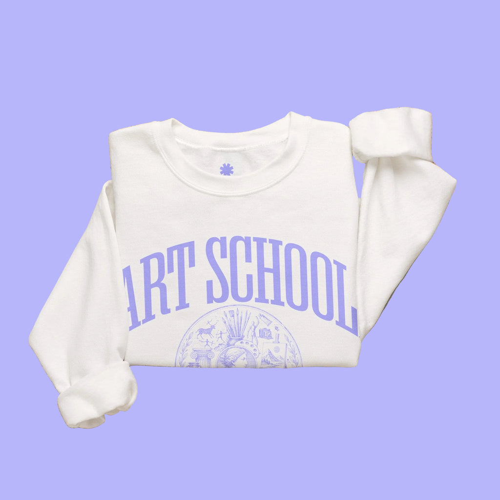 A folded white sweatshirt with purple 'ART SCHOOL' text and a circular graphic on the front, styled in a flat lay with woven textiles, a small candle jar, and a wicker tray on a light neutral background.