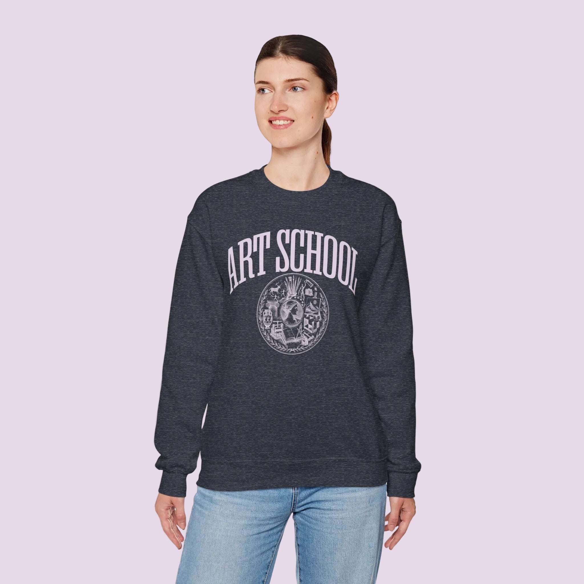 A woman modeling a dark heather gray crewneck sweatshirt with a distressed white graphic that reads 'ART SCHOOL' above a circular emblem, paired with light blue jeans against a plain white background.