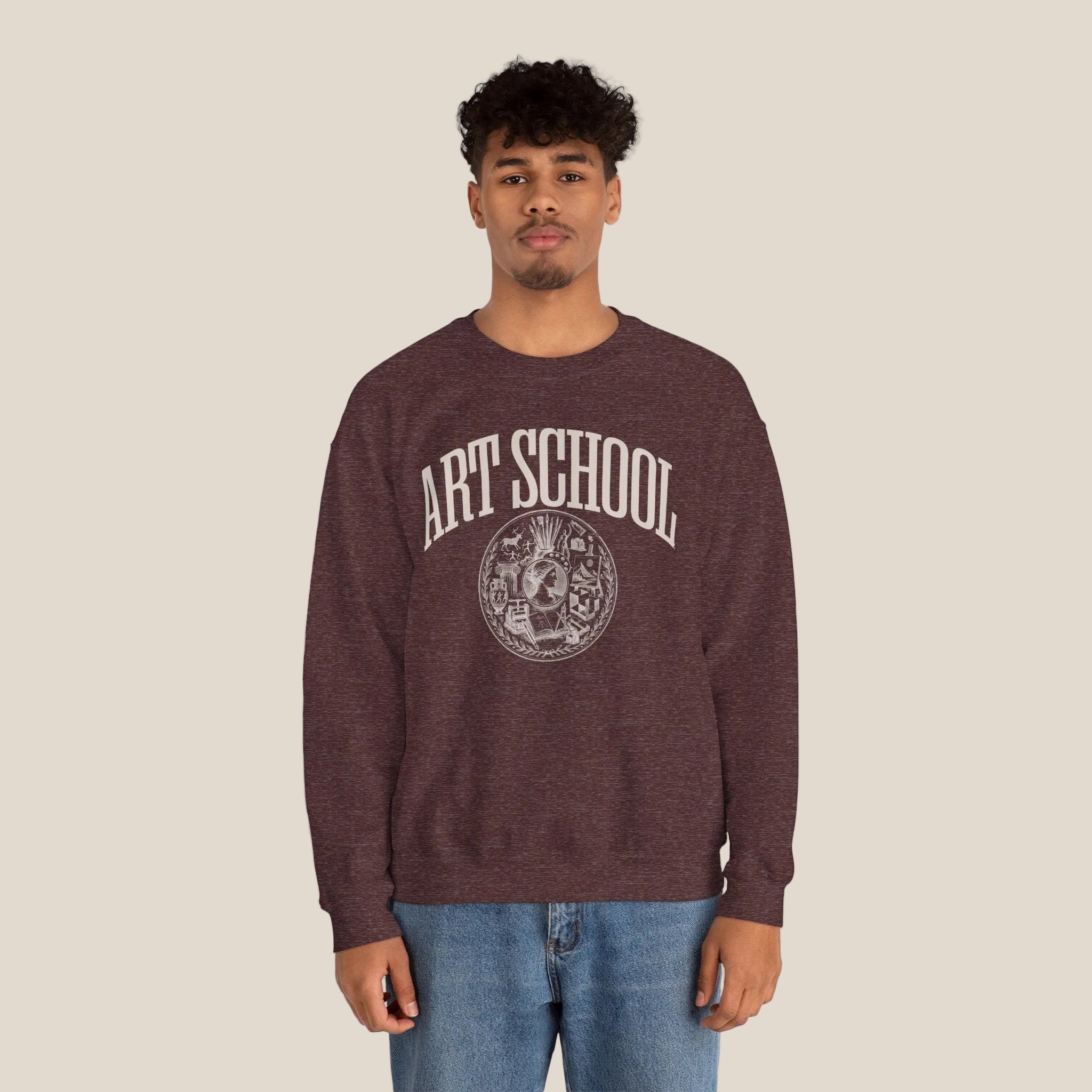 A man wearing a heathered dark brown crewneck sweatshirt with a distressed white graphic that reads 'ART SCHOOL' and a circular emblem, paired with blue jeans, against a plain white background.