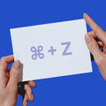 Two hands holding a white card with a lavender-colored 'COMMAND - Z' symbol against a solid blue background.