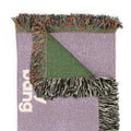 A folded woven throw blanket in lavender purple with a green triangular corner, multicolor fringe, and white woven lettering, shown on a plain white background.