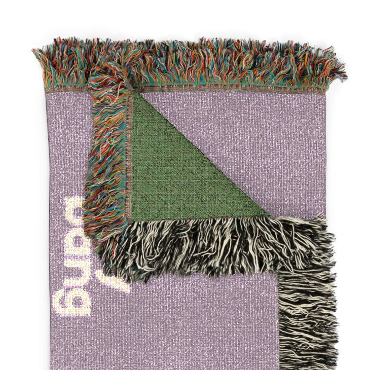 A folded woven throw blanket in lavender purple with a green triangular corner, multicolor fringe, and white woven lettering, shown on a plain white background.